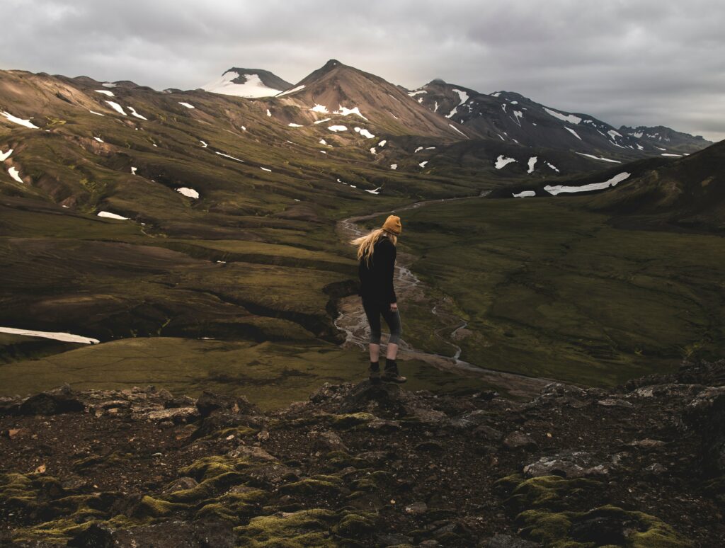 Girl in yellow beanie on mossy ridge overlooking green valleys and snow-capped mountains outdoors Wallpaper