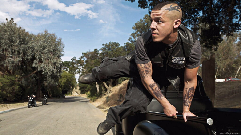 Pensive Rebel with Tattoos and Motorcycle Rider in Background – Ortiz Juice Wallpaper