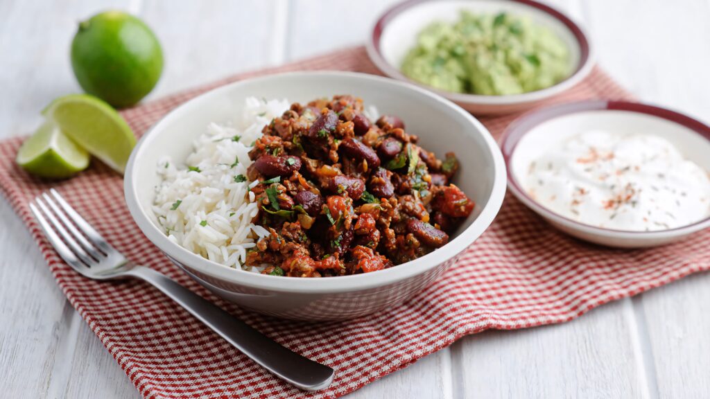 Hearty Chili Con Carne with beef beans rice guacamole sour cream lime on red checkered cloth wallpaper