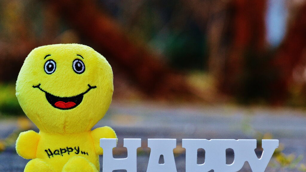 Cheerful plush yellow emoji doll with 3D white HAPPY letters on soft outdoor wallpaper background