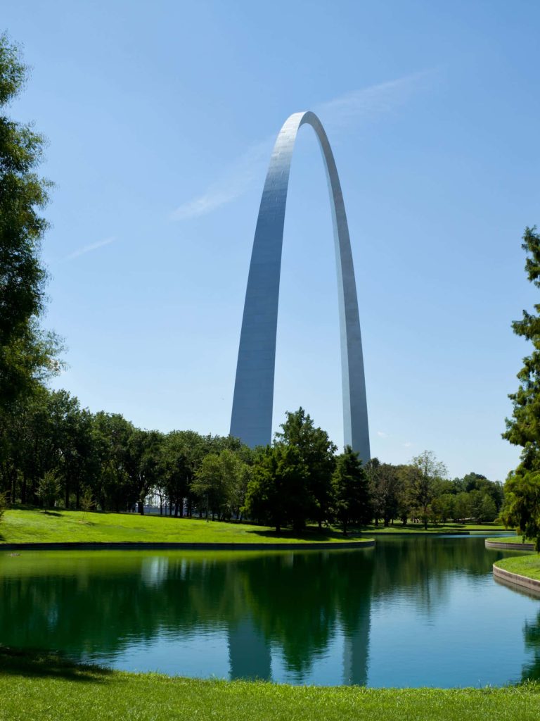 Gateway Arch in St Louis reflected in pond surrounded by green trees under clear blue sky Wallpaper