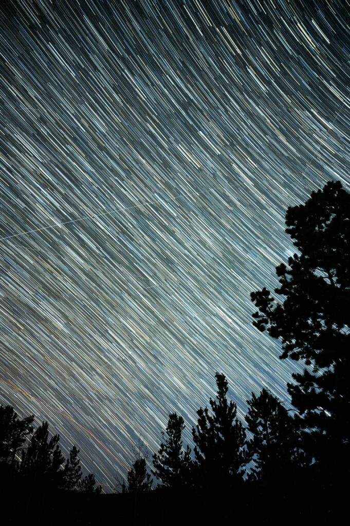 Starry sky wallpaper with dynamic star trails over dark forest silhouettes and vibrant night light stripes