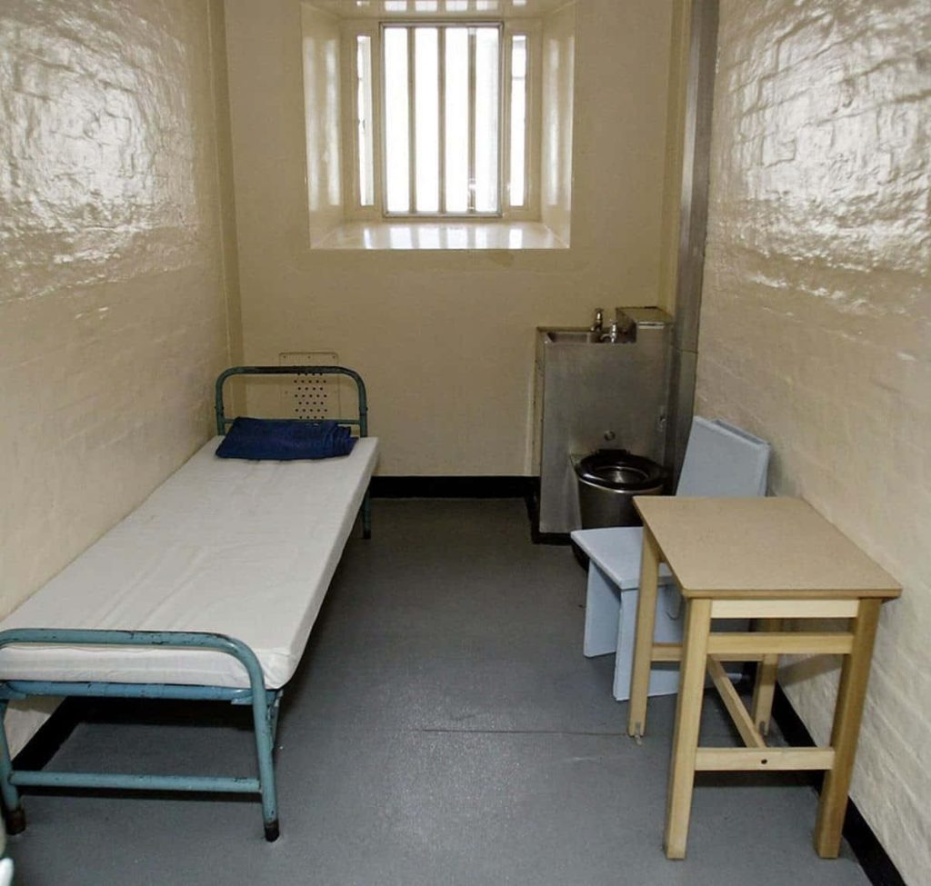 Stark prison cell with blue blanket cot metal sink-toilet and barred window brightly lit interior Wallpaper