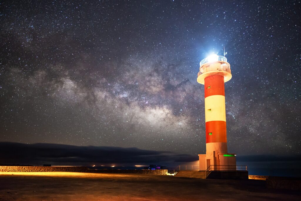 Striking red and white lighthouse under a vivid starry sky with Milky Way over coastal shore background Wallpaper