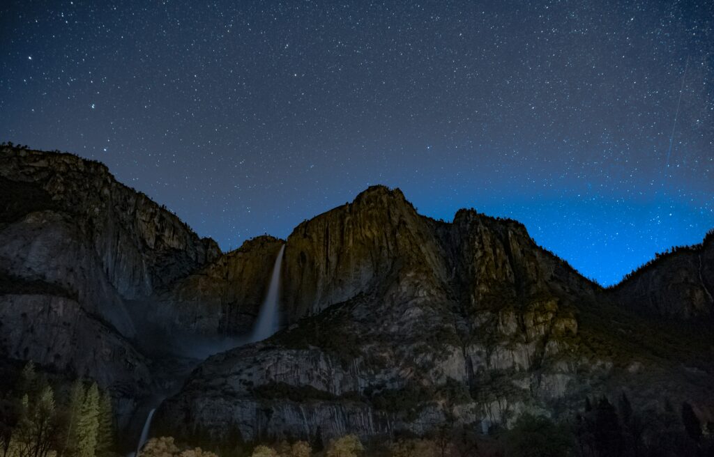 Starry night sky with a graceful waterfall over rocky cliffs in deep blues and moonlight Wallpaper