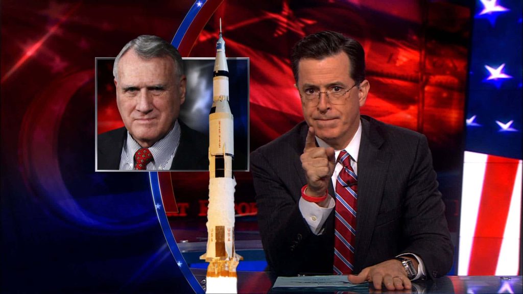 Stephen Colbert points from desk on The Colbert Report with Saturn V rocket and Jon Kyl graphic Wallpaper
