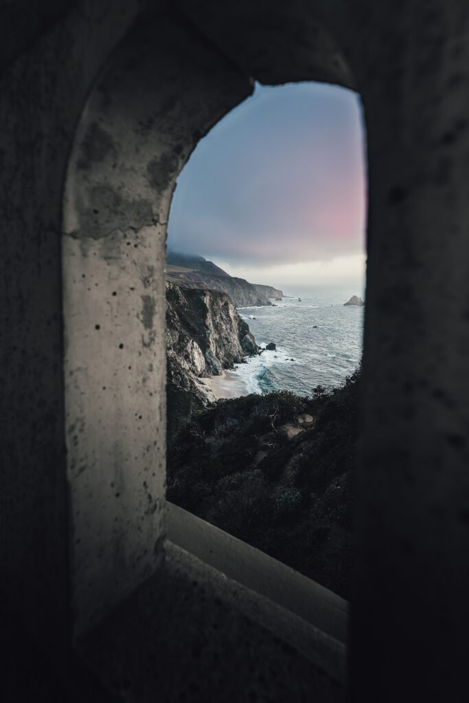 Coastal landscape through stone arch with rugged cliffs shoreline and calm sea under soft sky Wallpaper