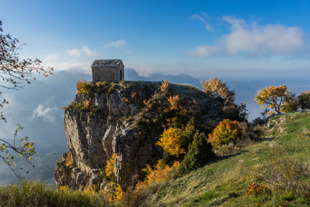 Small stone house on rocky cliff surrounded by autumn trees misty mountains blue sky wallpaper