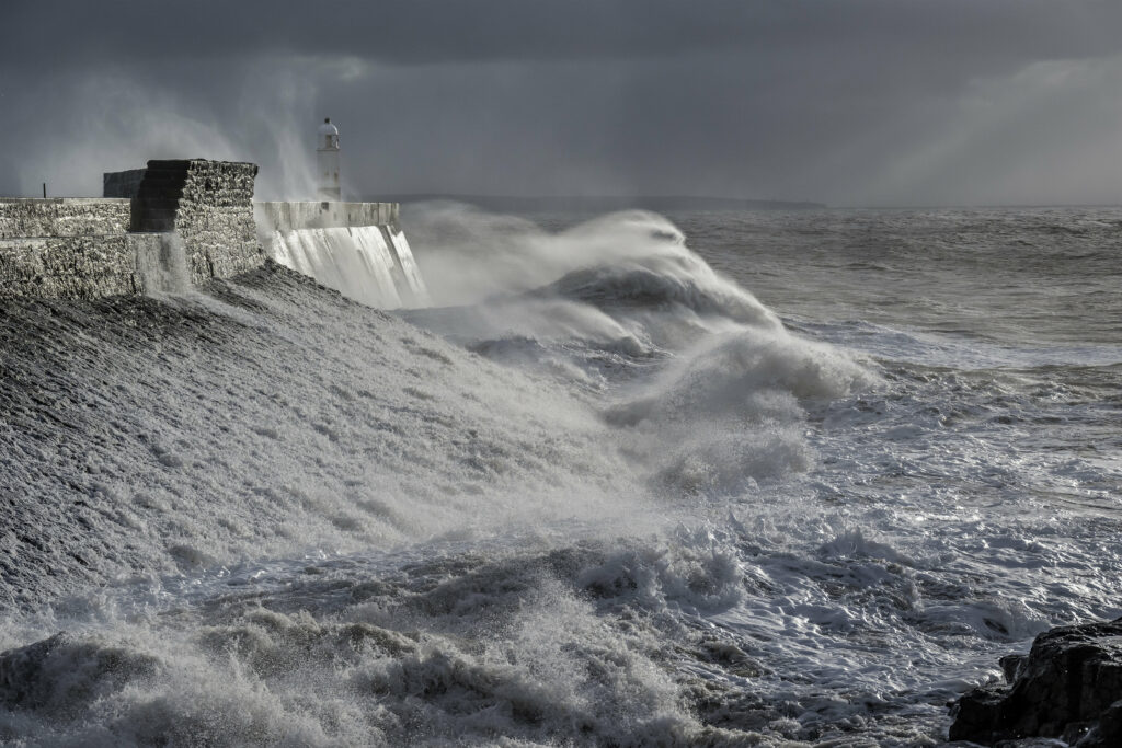 Stormy sea with foamy waves crashing on rocky shore and lighthouse in moody gray tones wallpaper