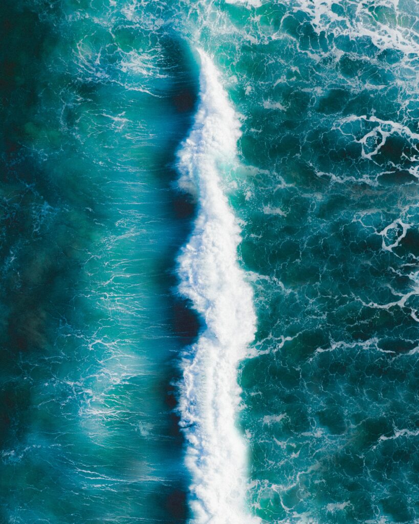 Aerial view of vibrant teal ocean wave crashing with dynamic white foam and swirling currents background Wallpaper