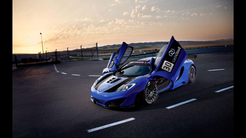 Blue McLaren with iconic doors open on racetrack at sunset showcasing luxury and performance Wallpaper