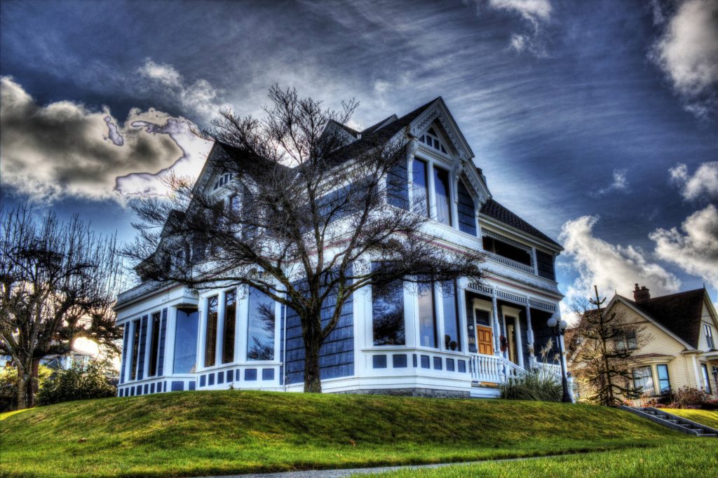 Blue Victorian house wallpaper with white trim dramatic sky and bare tree on grassy hill background