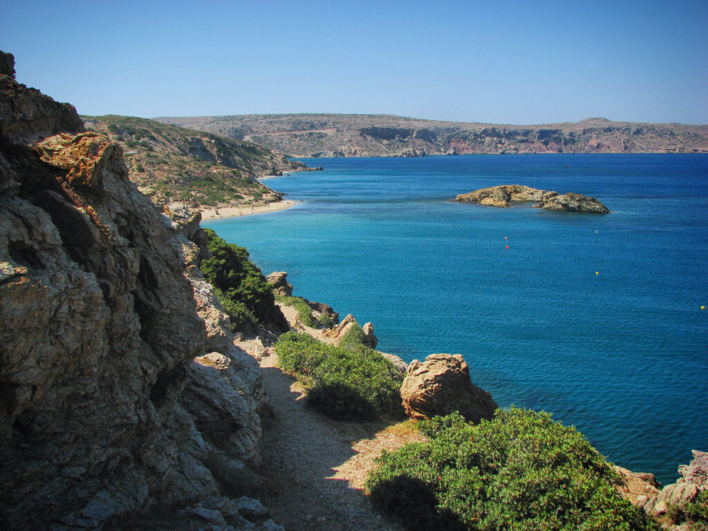 Coastal rocky cliffs with lush greenery meeting deep blue sea and distant mountains under clear sky Wallpaper