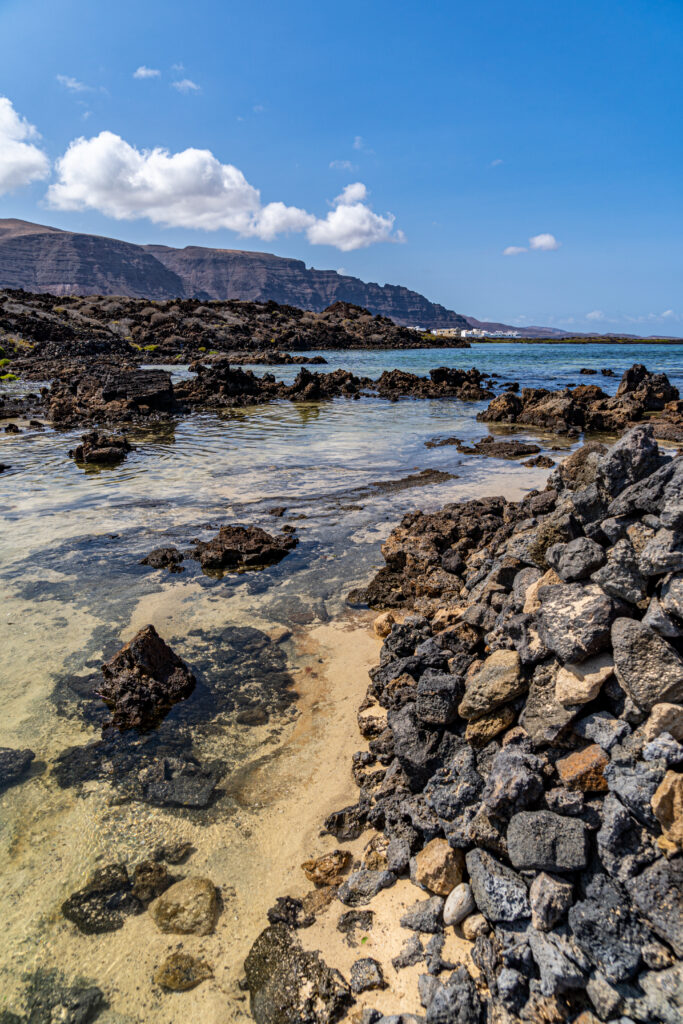 Coastal landscape with black stones clear shallow water mountains blue sky and fluffy clouds wallpaper