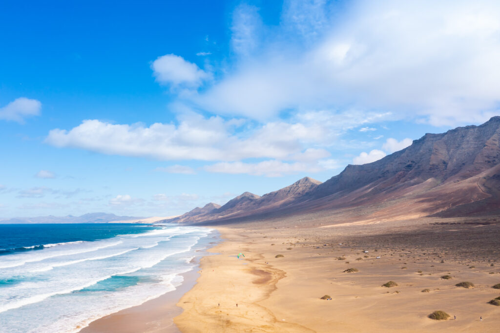 Coastal landscape with golden sandy beach waves white foam rugged mountains and blue sky clouds Wallpaper