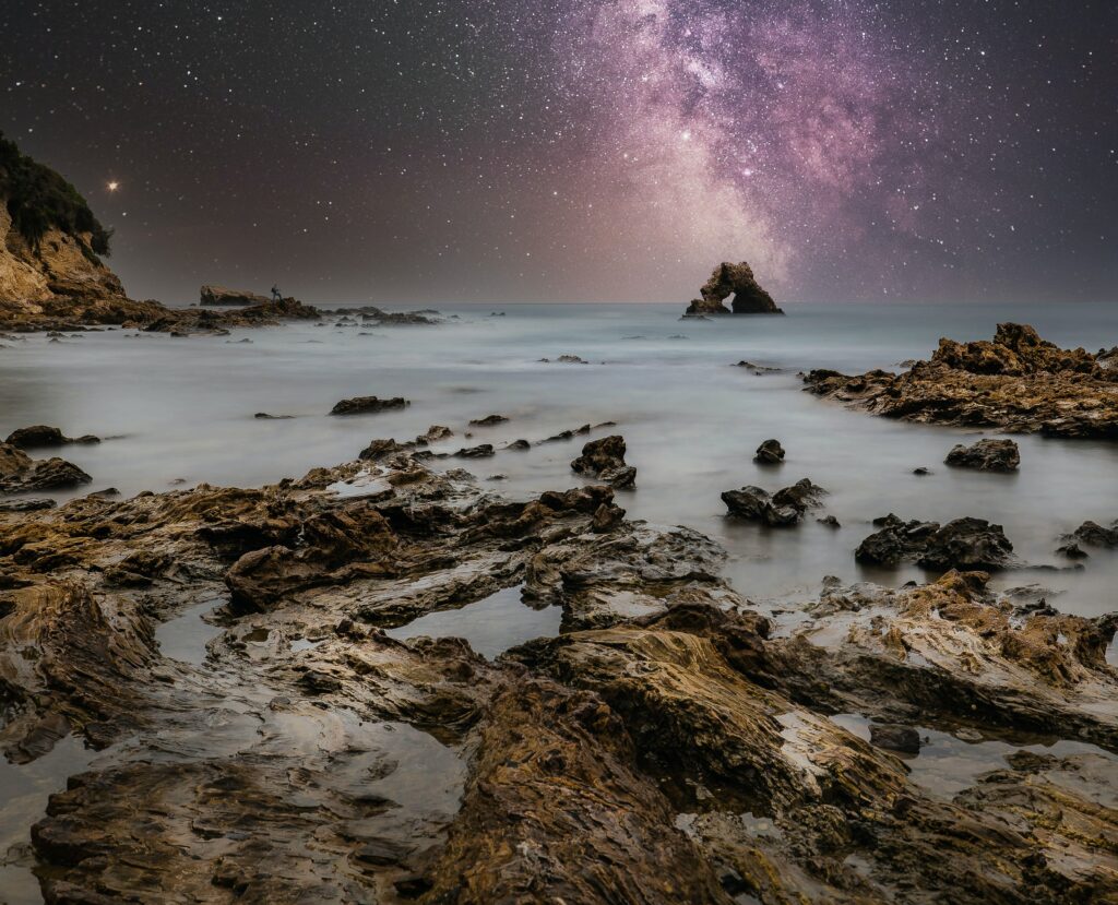 Starry night over rugged coastal rocks with serene water and vibrant Milky Way wallpaper