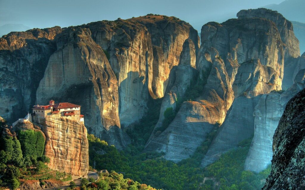 House atop massive rock formations surrounded by cliffs and green trees in warm sunlight view Wallpaper