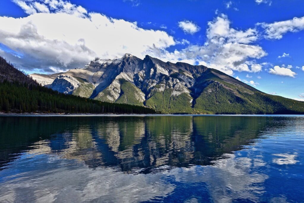 Mountain towering over calm lake with lush slopes reflected under blue sky and white clouds Wallpaper