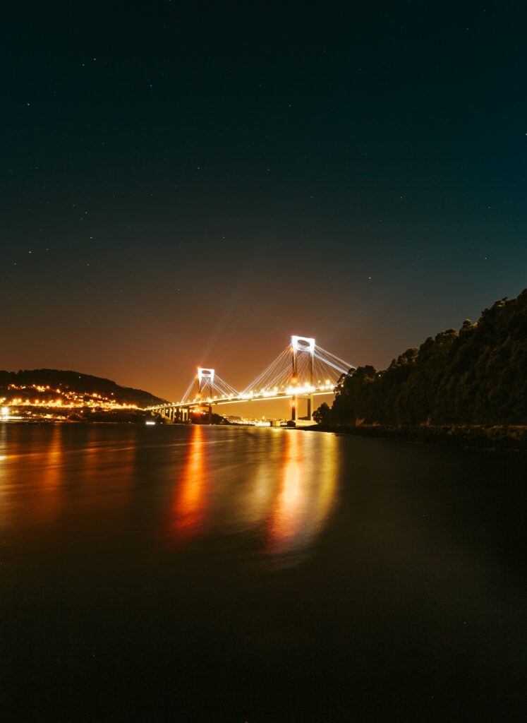 Night view of vibrant illuminated bridge reflecting on calm sea with hills and starry sky Wallpaper