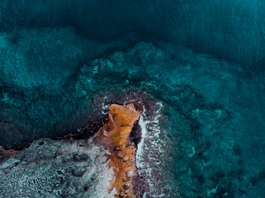 Top view of ocean waves swirling around rocky shore with sandy beach vibrant blue water wallpaper