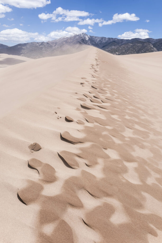 Soft sand dunes with wind patterns stretch toward rugged mountains under a bright blue sky wallpaper