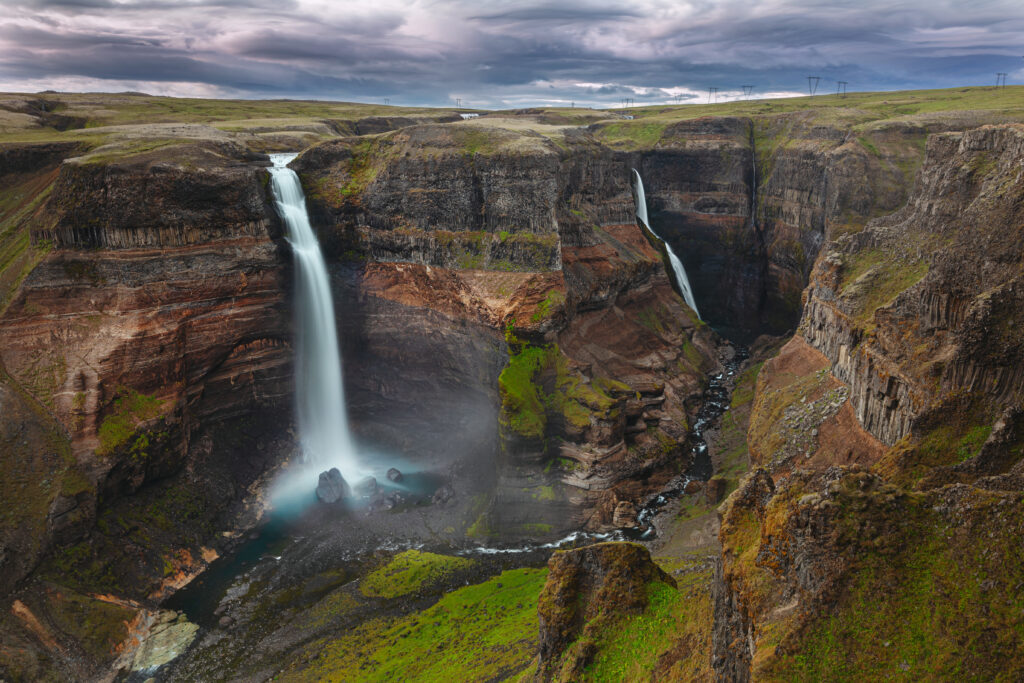 Waterfall cascading down rocky cliffs surrounded by lush green grass under dramatic cloudy sky Wallpaper
