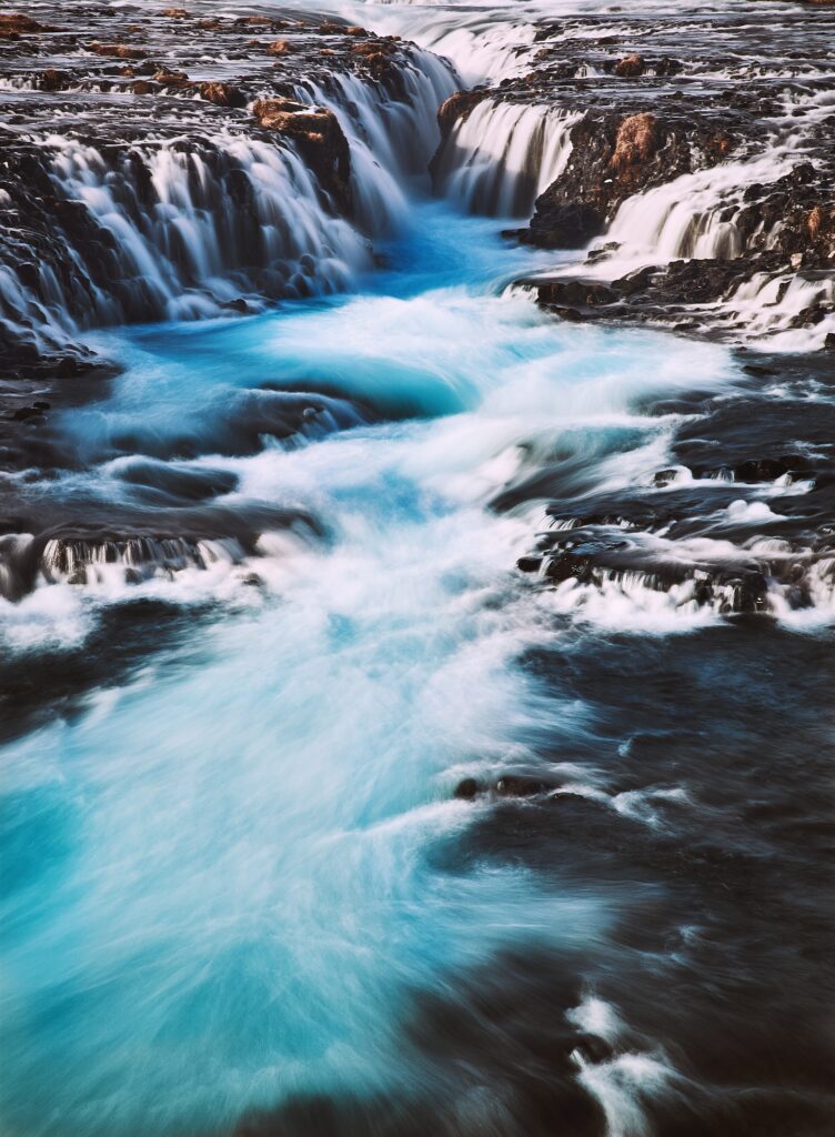 Waterfall cascading over dark rocks into vibrant blue river natural landscape wallpaper background