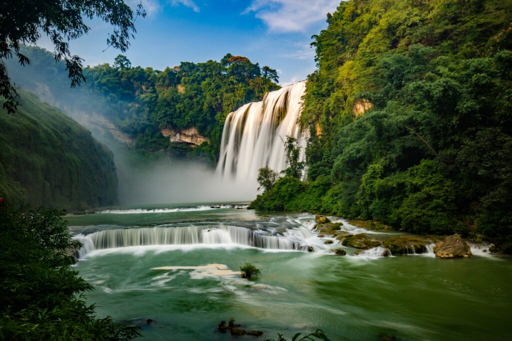 Waterfall cascading into vibrant river surrounded by lush green trees and flowing currents Wallpaper
