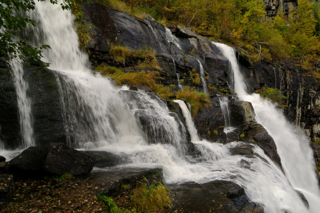Waterfall cascading over dark rocks with lush green grass and vibrant autumn foliage background Wallpaper