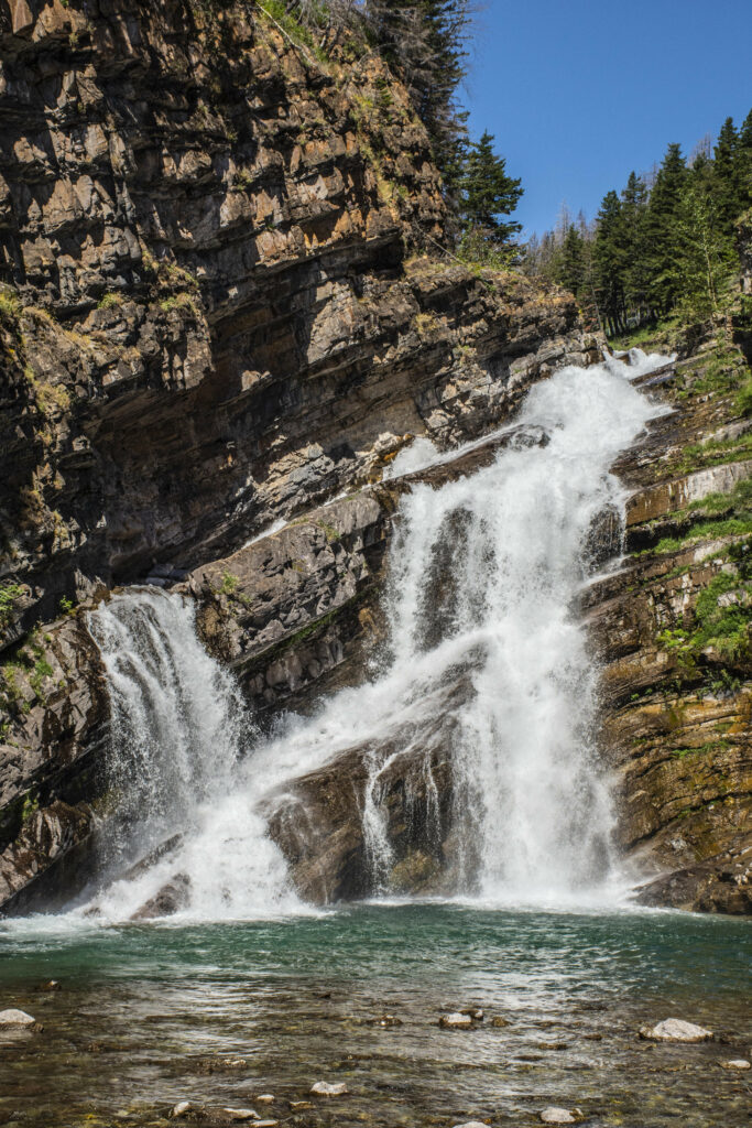 Stunning waterfall cascading over rugged rocks into clear pool with cliffs and green foliage Wallpaper