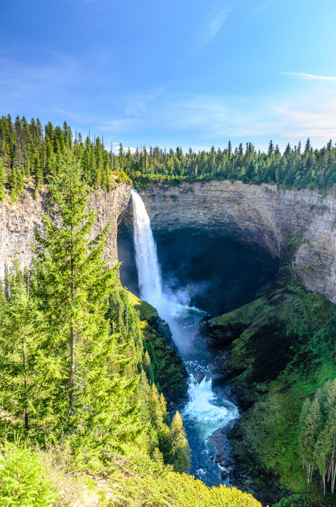 Waterfall cascading over rocks with spruce trees and vibrant greenery under a bright blue sky Wallpaper