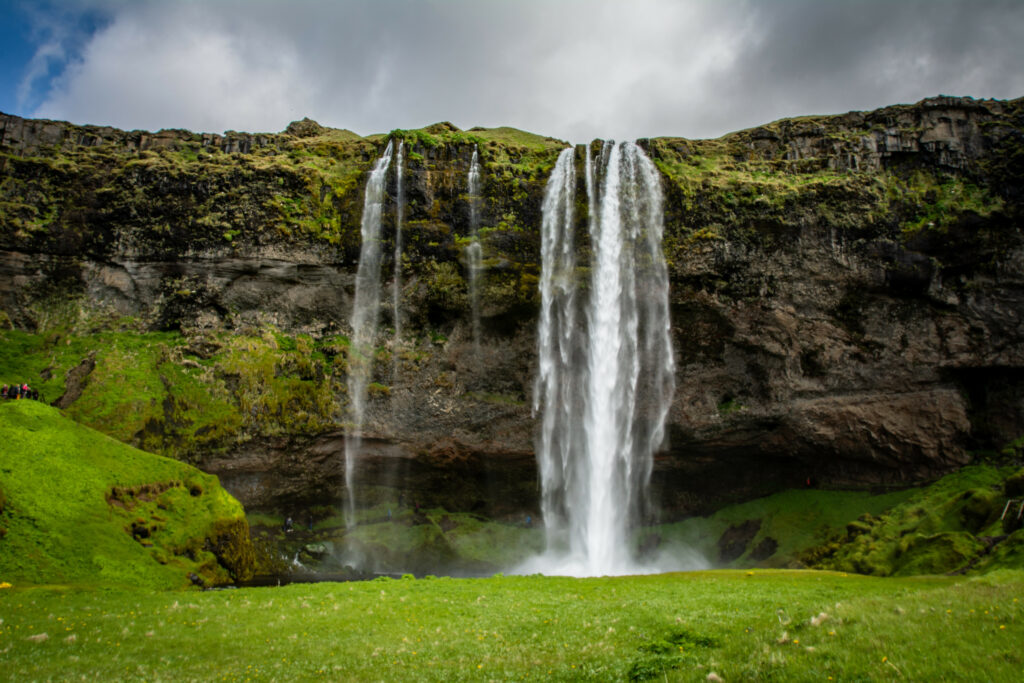 Waterfall cascading down rugged cliff with lush green grass moss and dramatic cloudy sky background Wallpaper