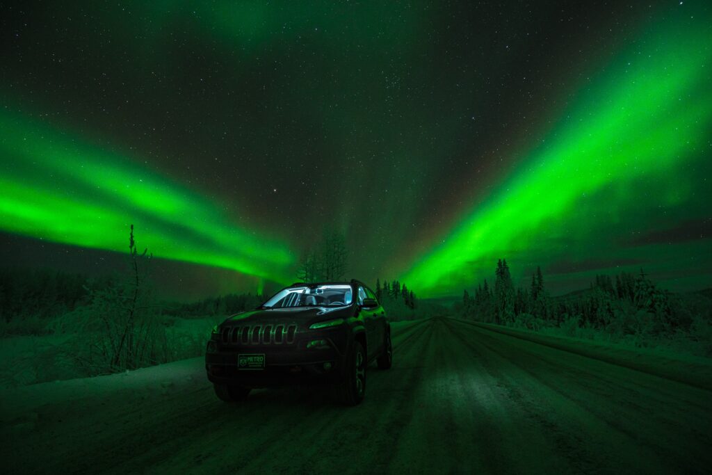 Winter Wonderland: Car on Snowy Road Under Northern Lights Wallpaper