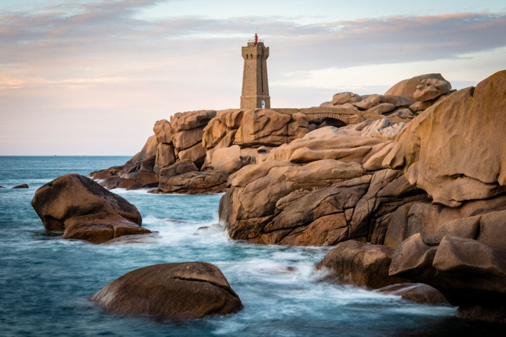 Sturdy lighthouse on rugged coastline with massive rocks and gentle waves under pastel sky wallpaper