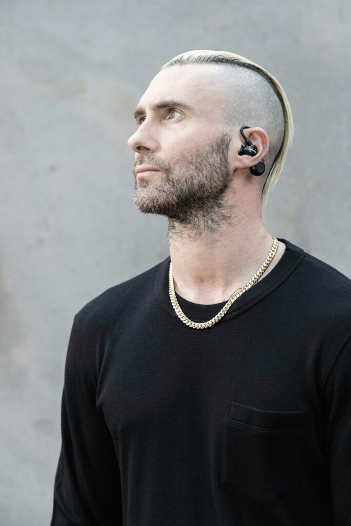 Stylish portrait of Adam Levine with slicked-back mohawk, black attire, and gold chain. Wallpaper