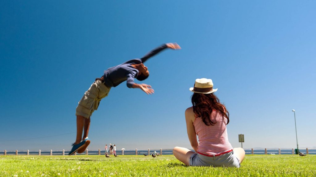 Man doing backflip on sunny green lawn near woman in straw hat under clear blue sky outdoors Wallpaper