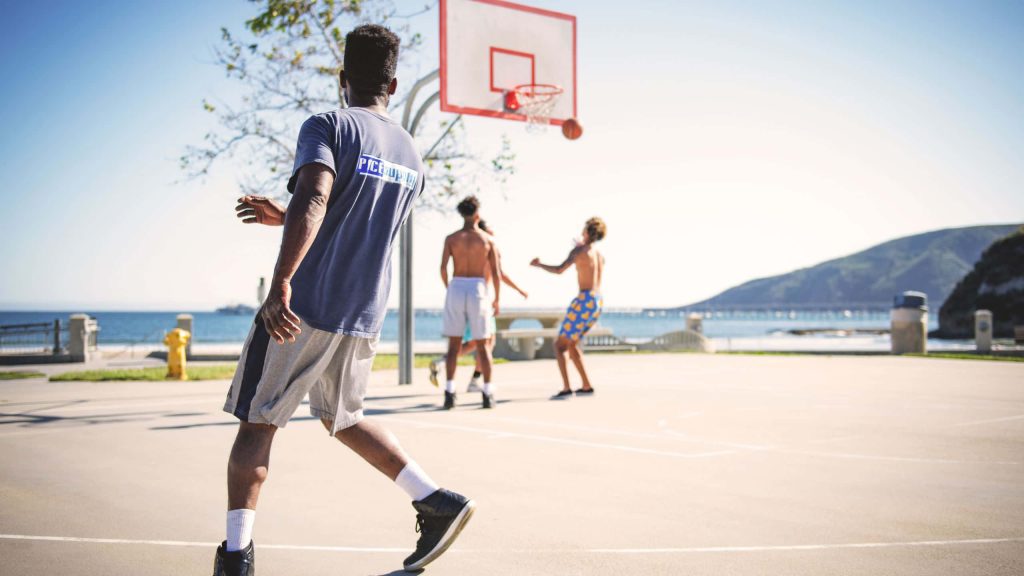 Men playing basketball on sunlit coastal court with ocean view HD sports wallpaper background