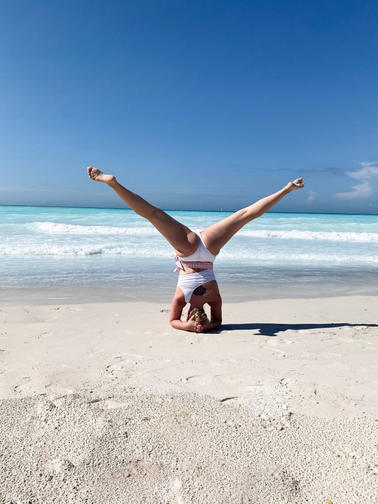 Woman in white swimsuit doing yoga inversion on white sand beach with turquoise water background Wallpaper