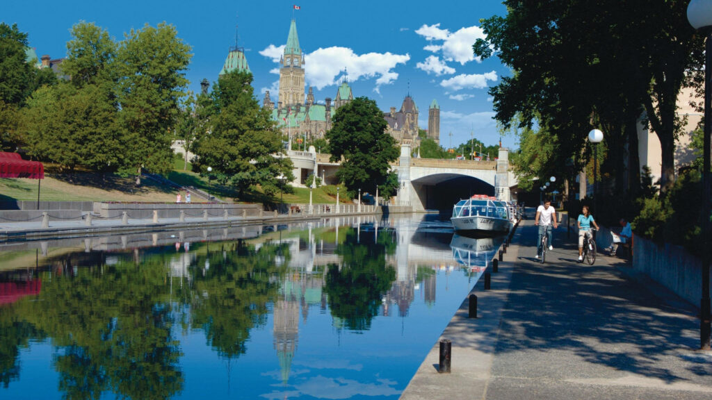 Vibrant summer Rideau Canal Ottawa with Parliament buildings biking walking lush trees blue water Wallpaper