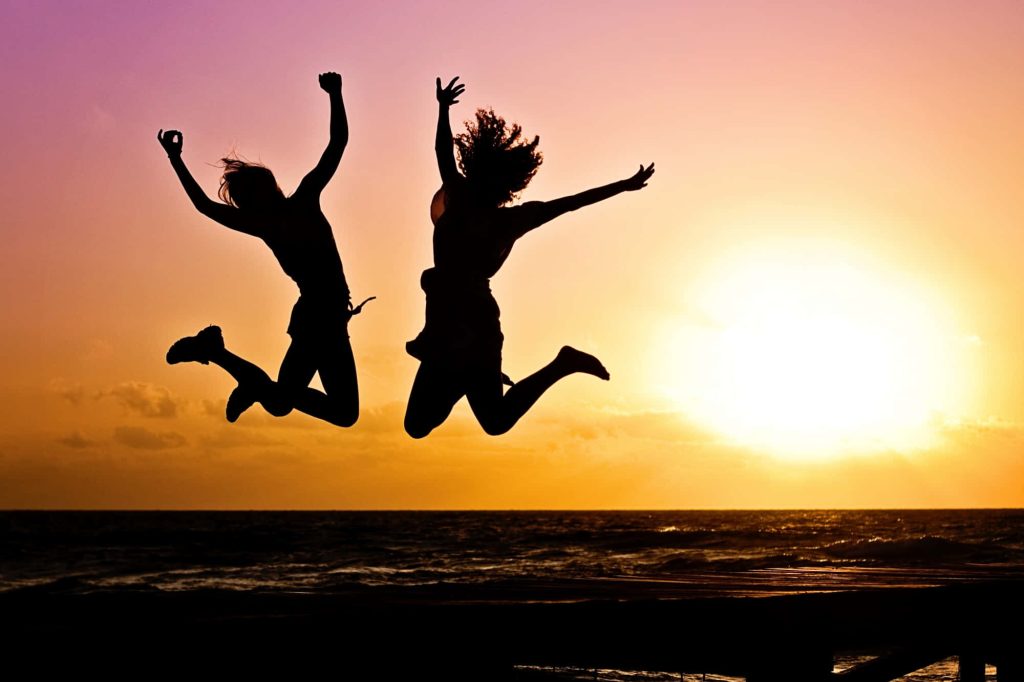 Silhouette of two people jumping at sunset over ocean vibrant summer wallpaper background