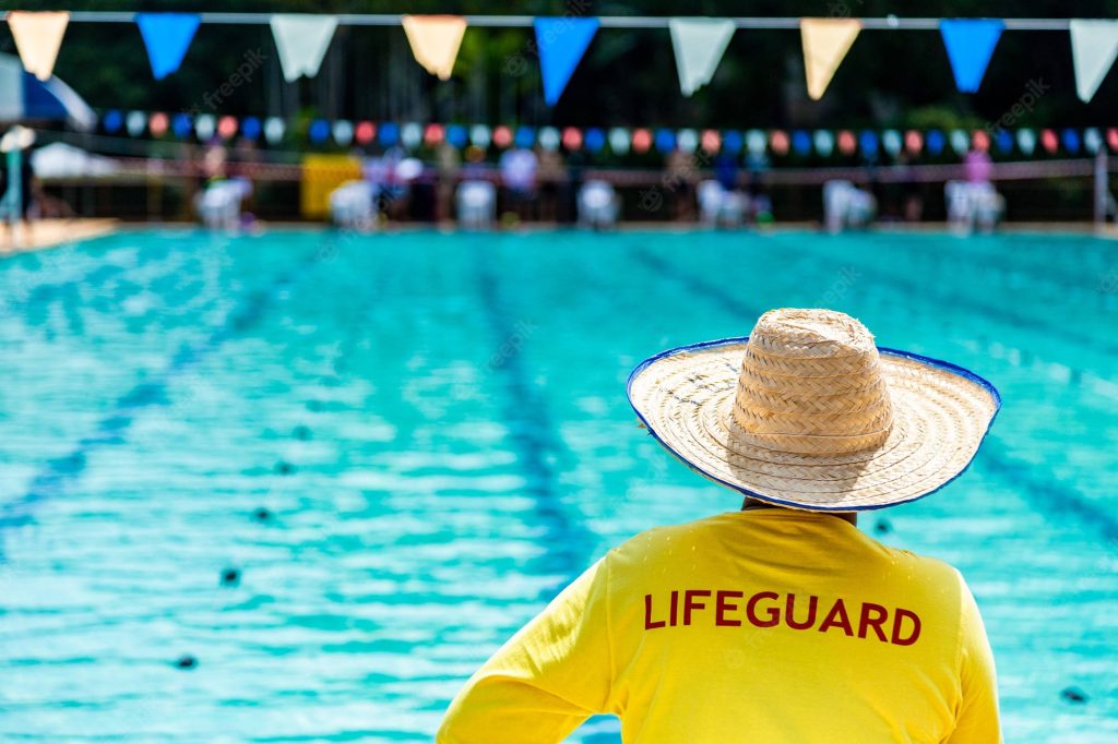 Lifeguard in yellow shirt and straw hat overlooking blue pool with colorful flags summer wallpaper