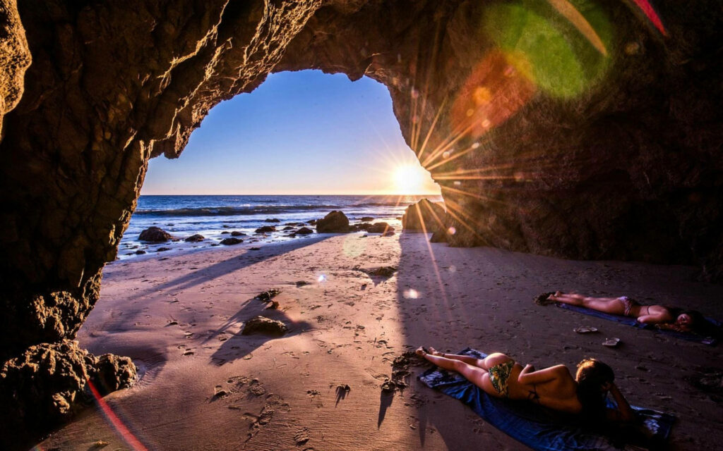 Secluded Serenity: Malibu Beach Retreat with Bikini-Clad Sunbathers Inside a Coastal Cave Embracing the Majestic Horizon Wallpaper