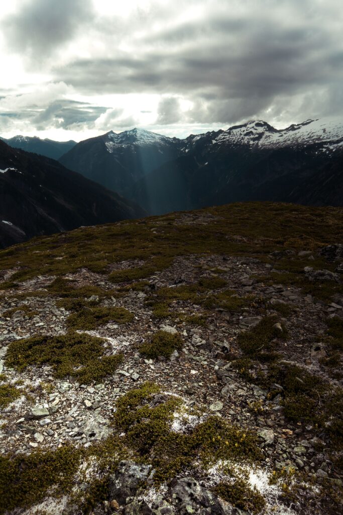 Sun rays break through clouds over rugged mountains with rocky terrain and distant snow peaks Wallpaper