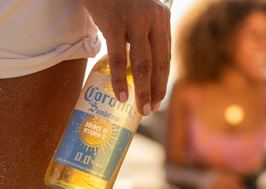 Close-up of hand holding Corona Sunbrew bottle on sunny beach with warm golden tones summer vibe Wallpaper