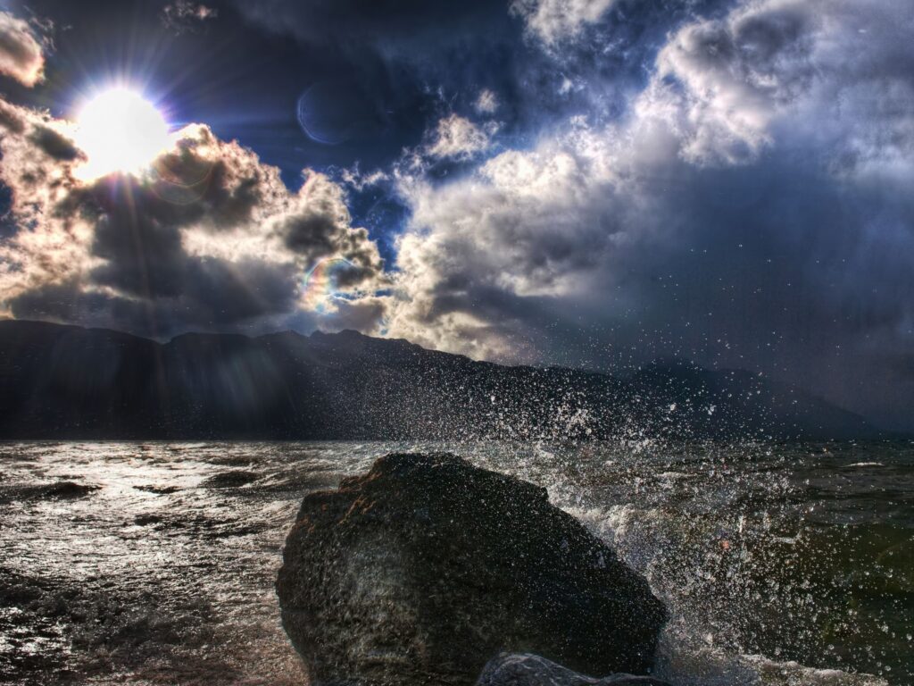 Sunlight breaks through clouds lighting rocky shore with splashing water under moody sky wallpaper