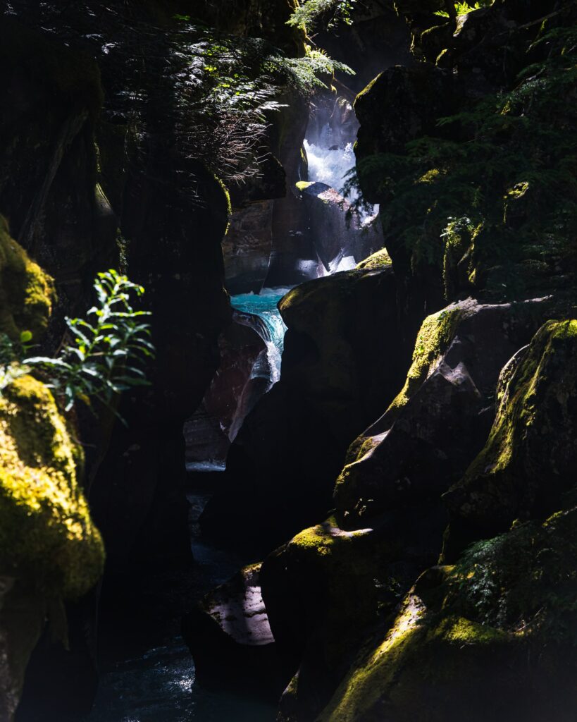 Sunlight shines on mossy rocks and vibrant waterfall flowing into deep blue gorge with lush branches Wallpaper