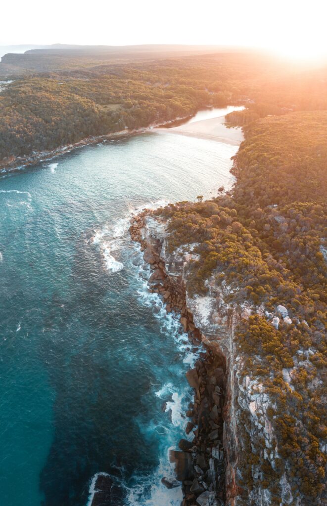 Aerial view of sunlit bay with clear blue sea cliffs and lush green trees coastal wallpaper background