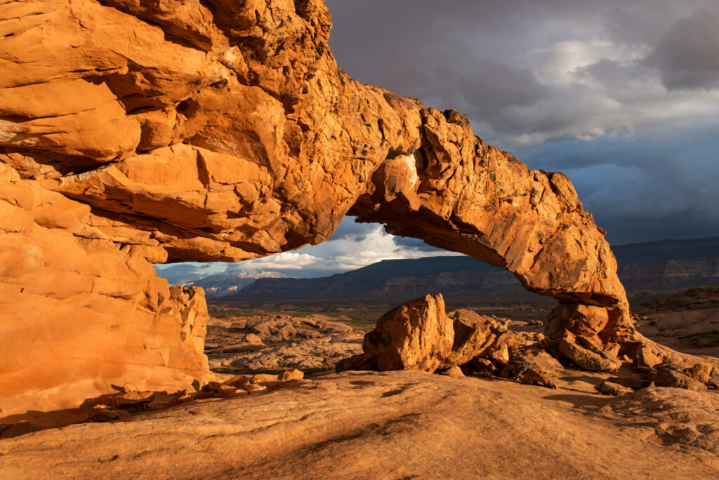 Sunlit natural rock arch in orange desert canyon with rugged formations and dramatic sky Wallpaper