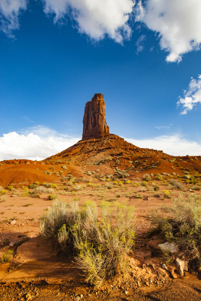 Striking rock formation in sunlit reddish prairie with rugged bushes under vivid blue sky clouds Wallpaper