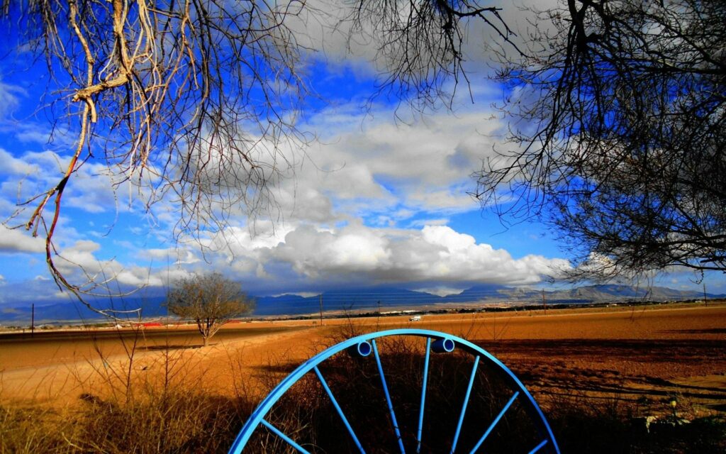 Rustic blue iron wheel in sunlit field under dramatic clouds and deep blue sky with bare trees Wallpaper